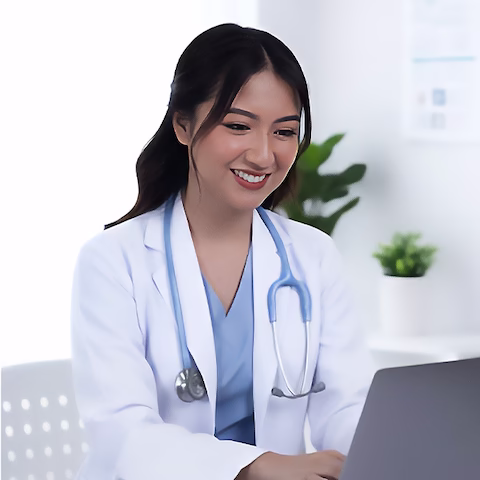 Dr. Maria Beatrice in a white coat and stethoscope smiling while using a laptop in a modern clinic office.